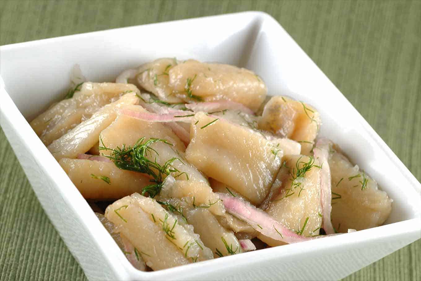 porcja śledzi w occie z dodatkami ziołowymi porcja marynowanych śledzi obok cytryny, oliwy i świeżych ziół na desce