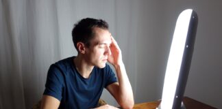 Terapia światłem w depresji sezonowej i nawracającej – jak codzienna dawka jasnego światła przywraca równowagę serotoniny i melatoniny? młoda kobieta siedząca przed jasną lampą terapeutyczną w przytulnym pokoju z kubkiem kawy