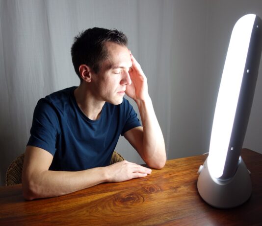 Terapia światłem w depresji sezonowej i nawracającej – jak codzienna dawka jasnego światła przywraca równowagę serotoniny i melatoniny? młoda kobieta siedząca przed jasną lampą terapeutyczną w przytulnym pokoju z kubkiem kawy